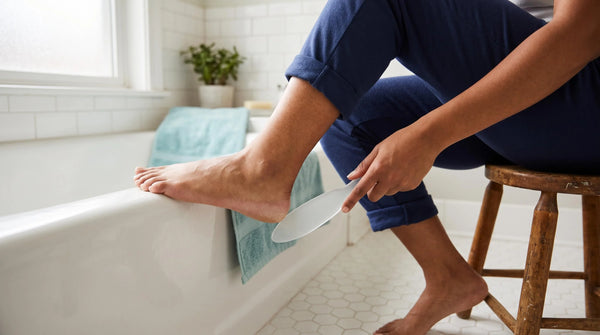 Woman sitting on a stool in her bathroom with her foot on the tub edge, using a glass foot file as a callus remover on her heel