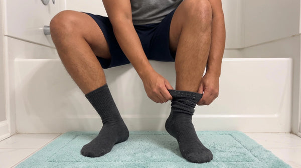 A man sitting on the edge of a bathtub pulling off a dark sock with lint on it, the kind of sock fuzz that turns into toe jam