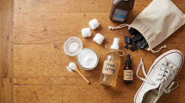 Flat lay of DIY shoe deodorizer ingredients on a wood surface: baking soda, alcohol, vinegar spray bottle, tea tree oil, charcoal pouch, and a white sneaker.