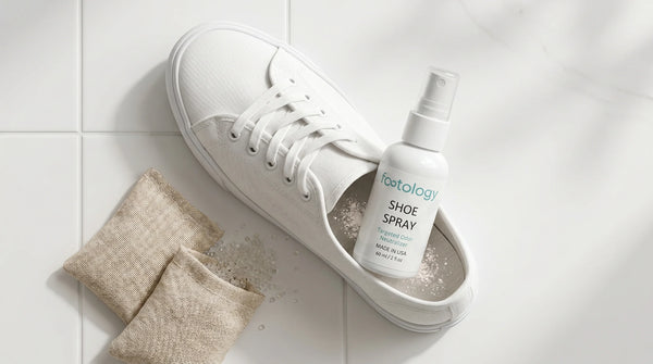 Flat lay of three shoe deodorizer formats on a white bathroom counter: aqua MyFootology shoe spray bottle, two natural fabric pouches with baking soda and charcoal, and a small powder dusting next to a white canvas sneaker.